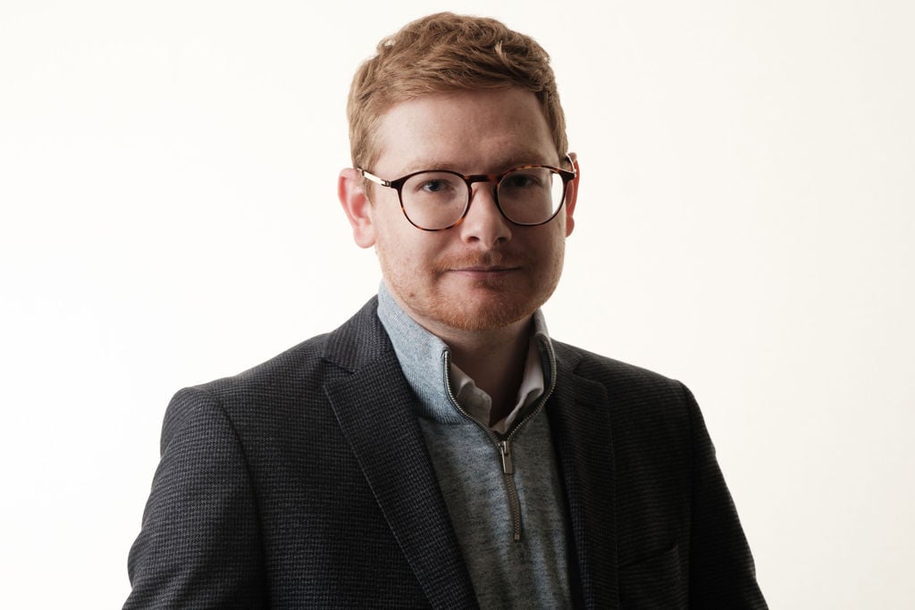 Tom Wardman in a dark blazer over a grey jumper, looking at the camera against a light background.