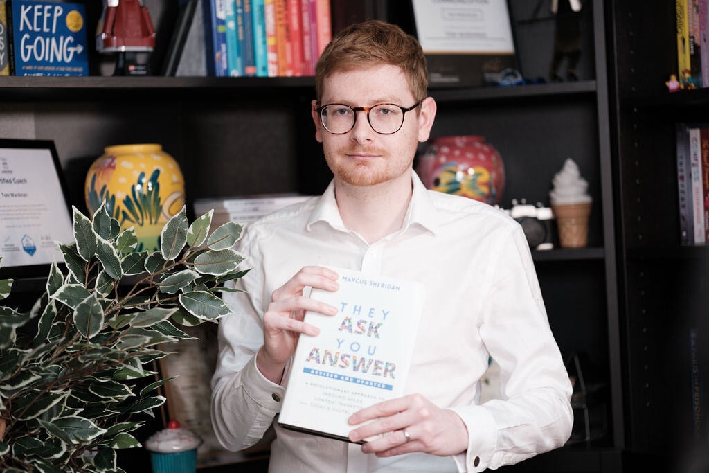 Tom Wardman holding a copy of They Ask, You Answer in front of a bookshelf.